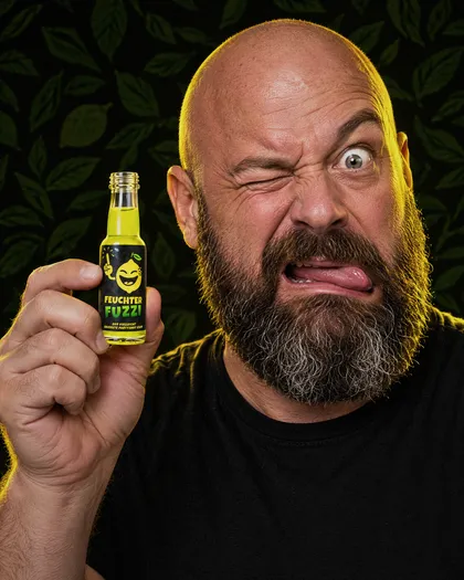 Bärtiger Mann mit Glatze hält ein Fläschchen, ein Auge zu, die Zunge draußen